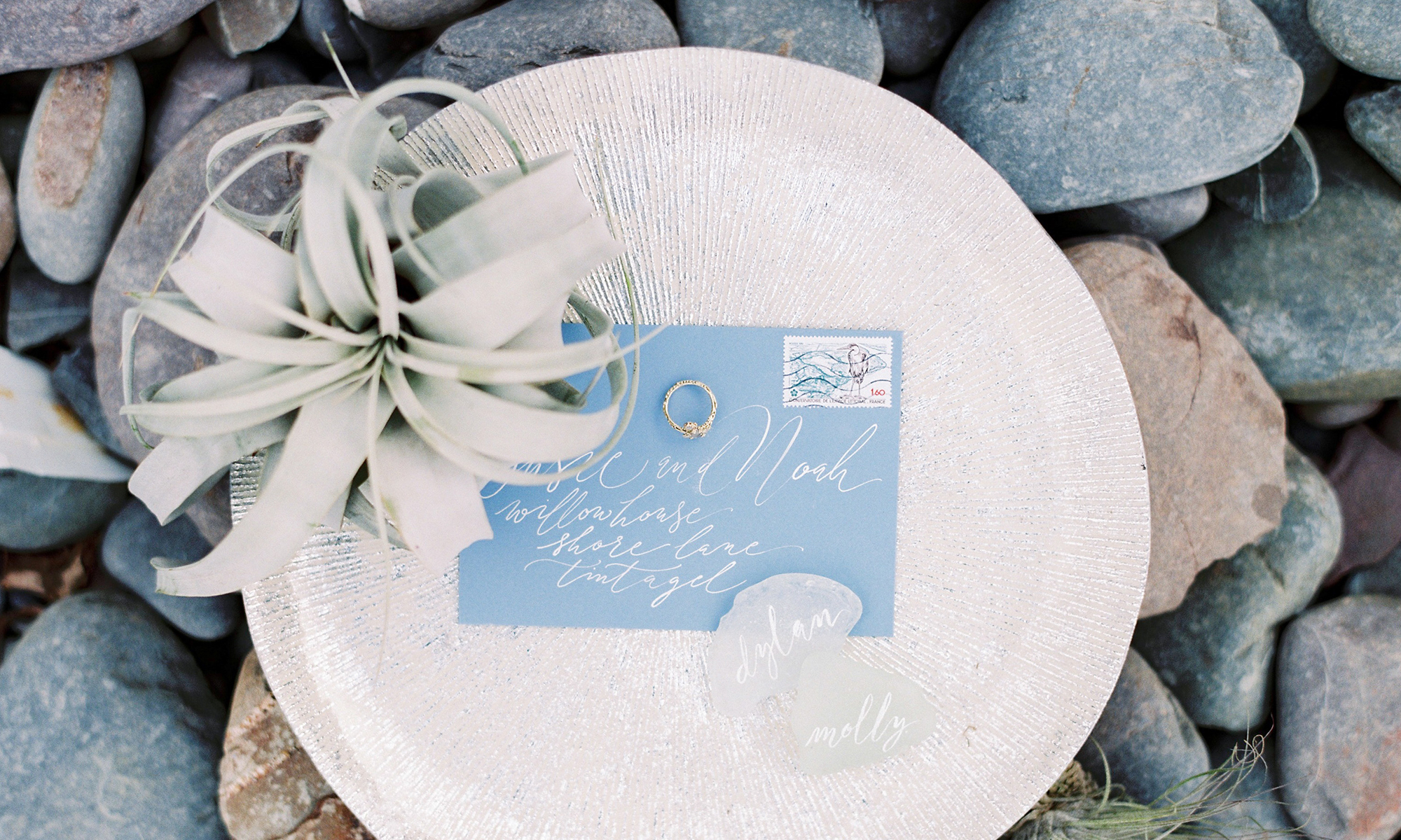 A pale blue envelope with calligraphy addressing on a silver plate with a succulent and sea glass, and a wedding ring. The plate is on pebbles, like on a beach