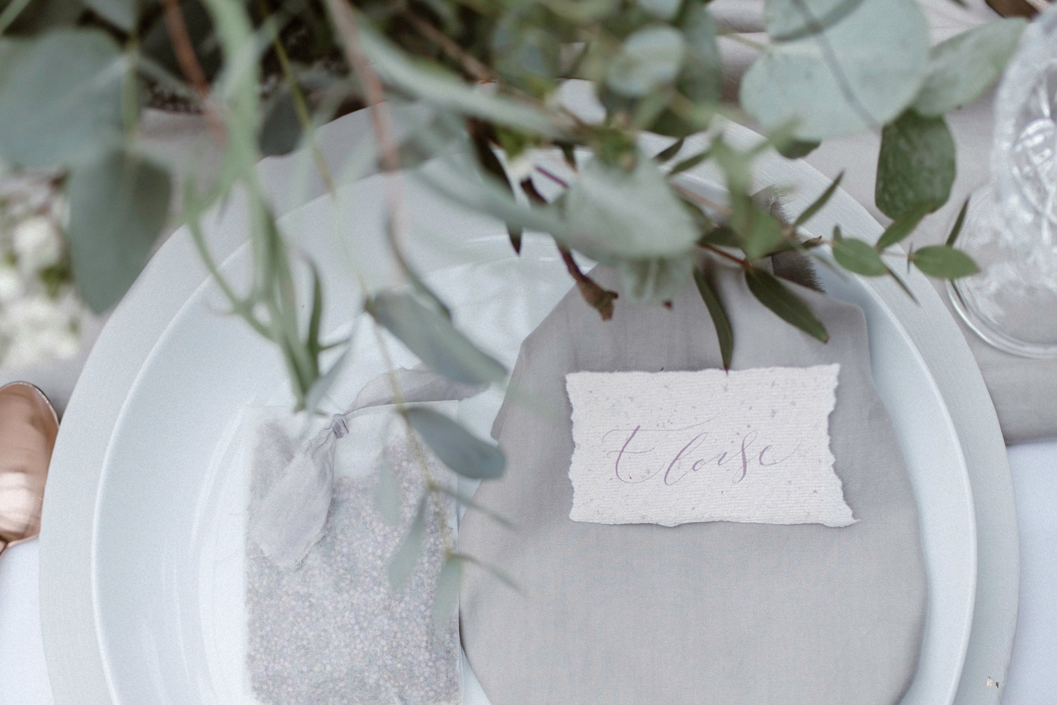 an elegant wedding place setting with a napkin and torn edge recycled calligraphy name card. Eucalyptus sprigs hang over the top of the plates, which are photographed from above