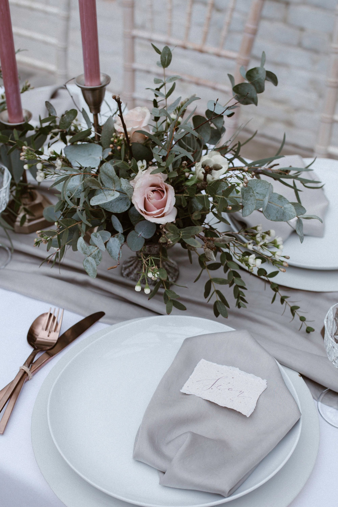 Wedding table with eucalyptus and pink roses, and dusky pink candles. White plates are styled simply with a grey napkin and calligraphy place name card