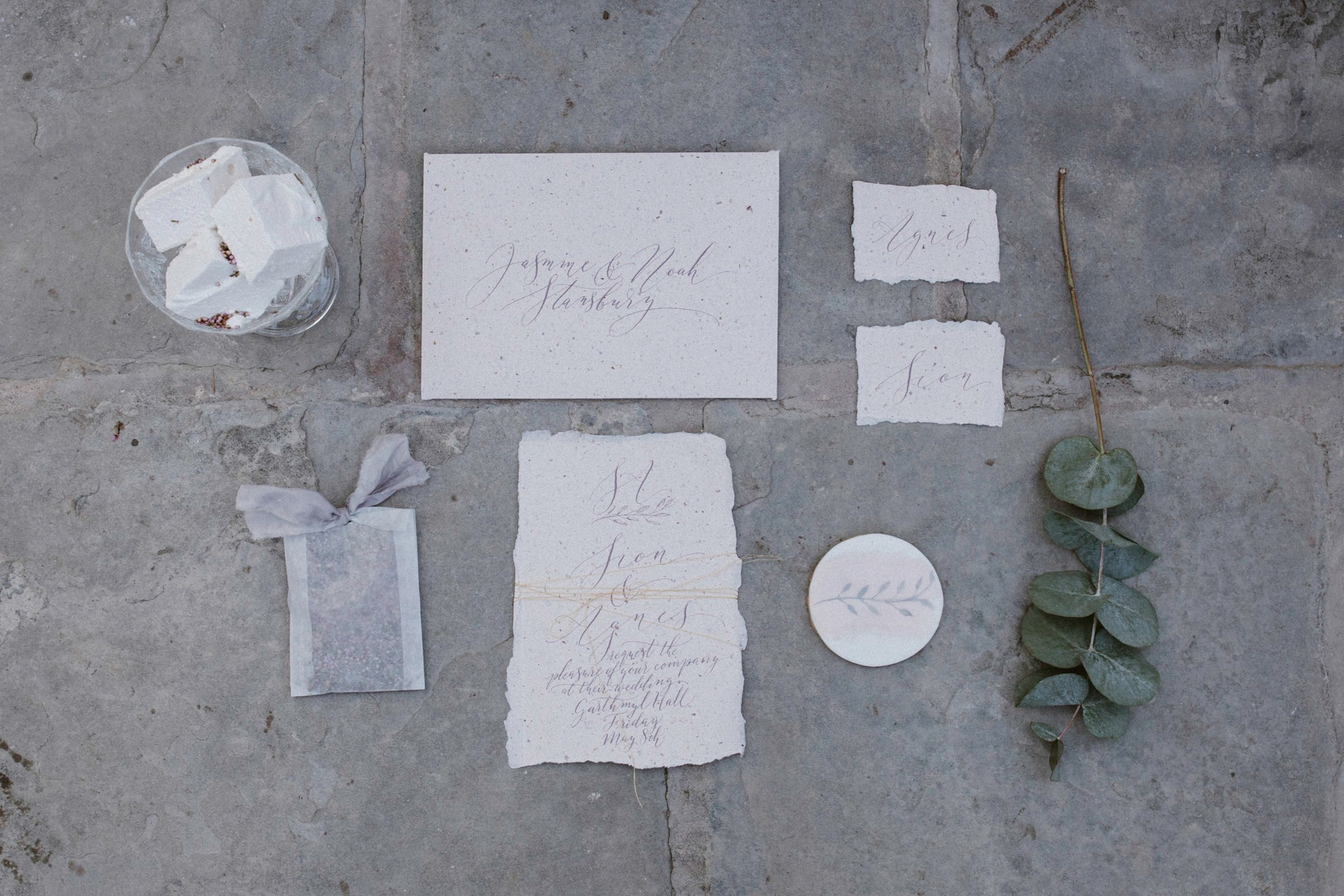 Chic minimal calligraphy for wedding stationery with an inner envelope and a torn edge invitation, and two calligraphy place names. These are on a stone floor alongside a eucalyptus spring, a handprinted cookie, a glassine bag of confetti and some artisan marshmallows