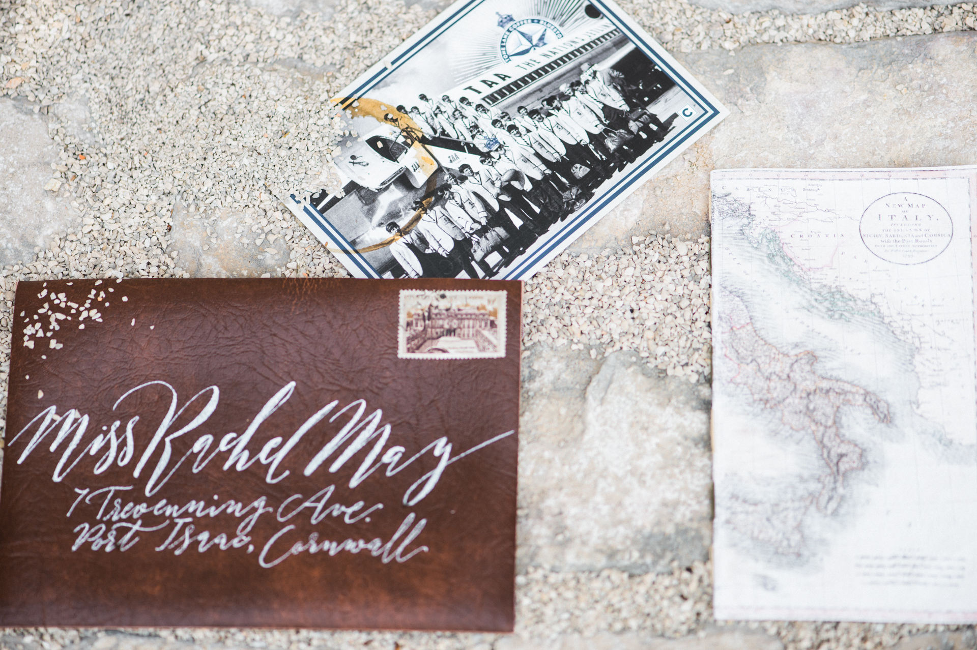 A faux leather envelope with handwritten modern calligraphy. The envelope is shown next to a vintage photo of men in suits lined up next to a plane, and a vintage map of Italy. Sand is scattered across the shot.