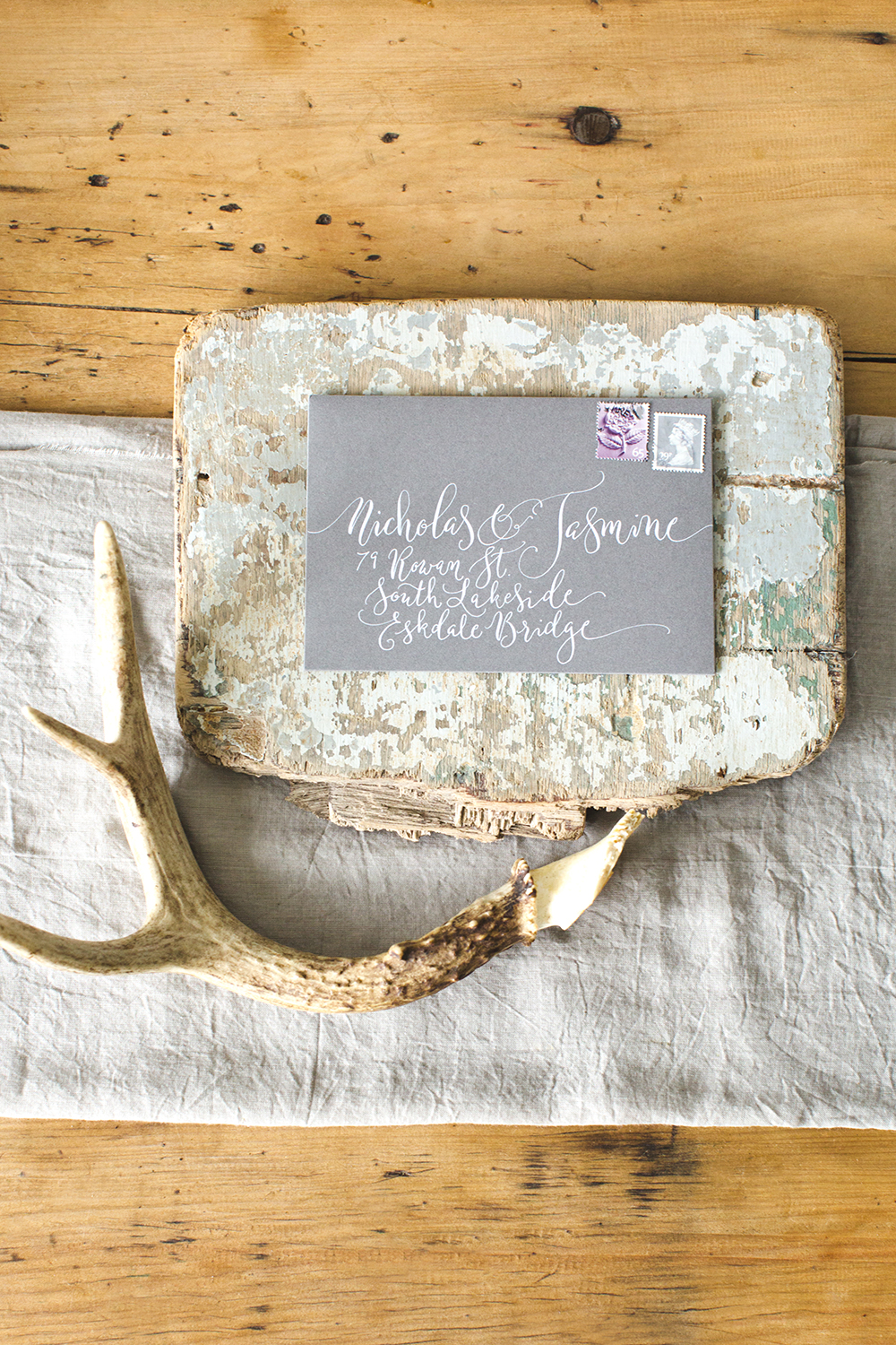 A grey calligraphy envelope with white lettering in a modern style sits on top of a scrap of old painted wood. There's a folded sheet of muslin underneath and a discarded deer antler to one side. 