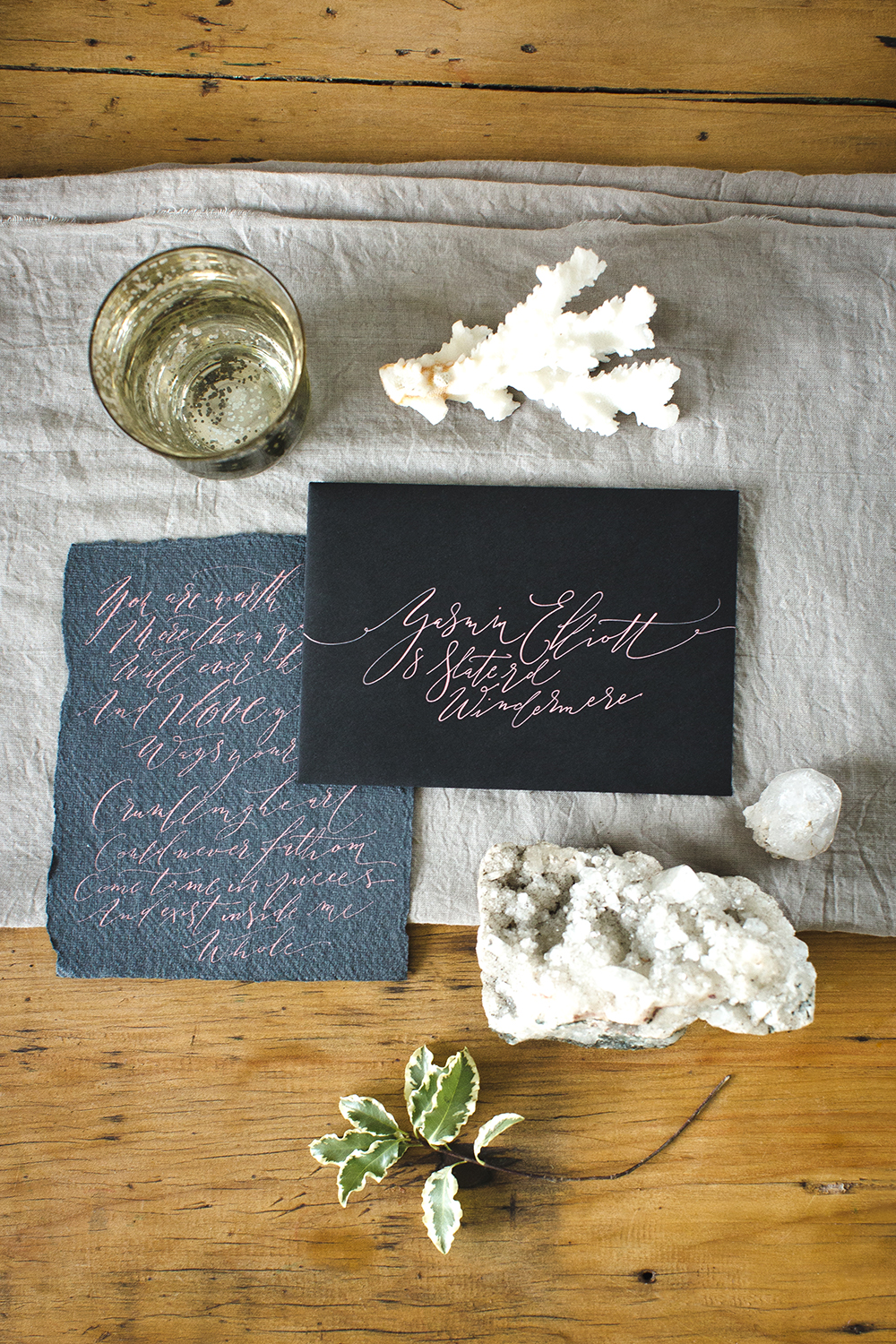 A calligraphy envelope with dusky pink ink on black paper, and a calligraphy love letter to one side. The handwritten wedding items are surrounded by natural objects including a chunk of quartz, some holly leaves and sea coral, and sat on a folded sheet of muslin cloth