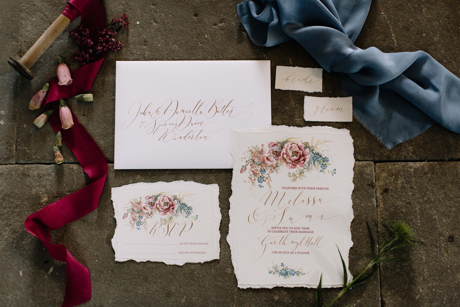 A wedding invitation, RSVP and calligraphy envelope in burgundy ink. The calligraphy is very modern and elongated. The invitation has a torn edge and floral illustrations. There are silk ribbons and flower buds scattered around, and a stone floor underneath
