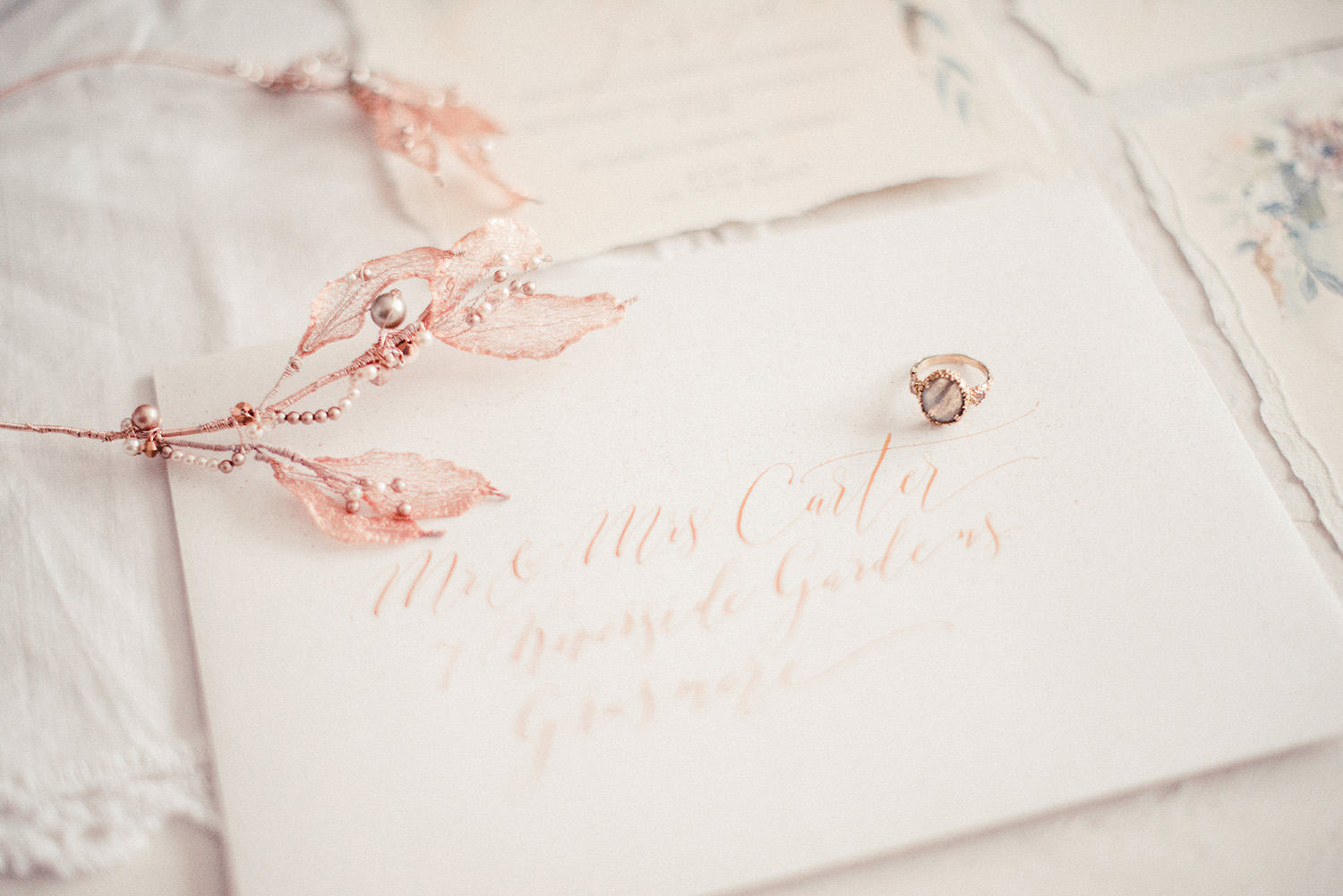 Pale rose gold calligraphy on a white envelope, with a delicate floral crown placed above, and a wedding ring on the envelope