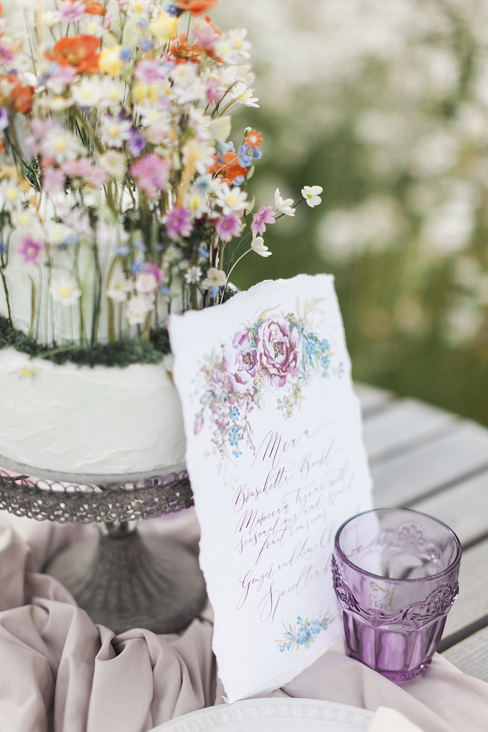 A colourful wedding cake covered in sugar paste wildflowers standing up like a meadow, and a wedding menu with floral illustration and hand calligraphy
