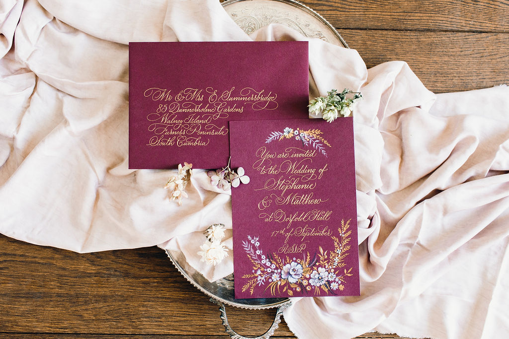 romantic modern wedding calligraphy envelope with swirling gold script on a burgundy envelope and floral illustration