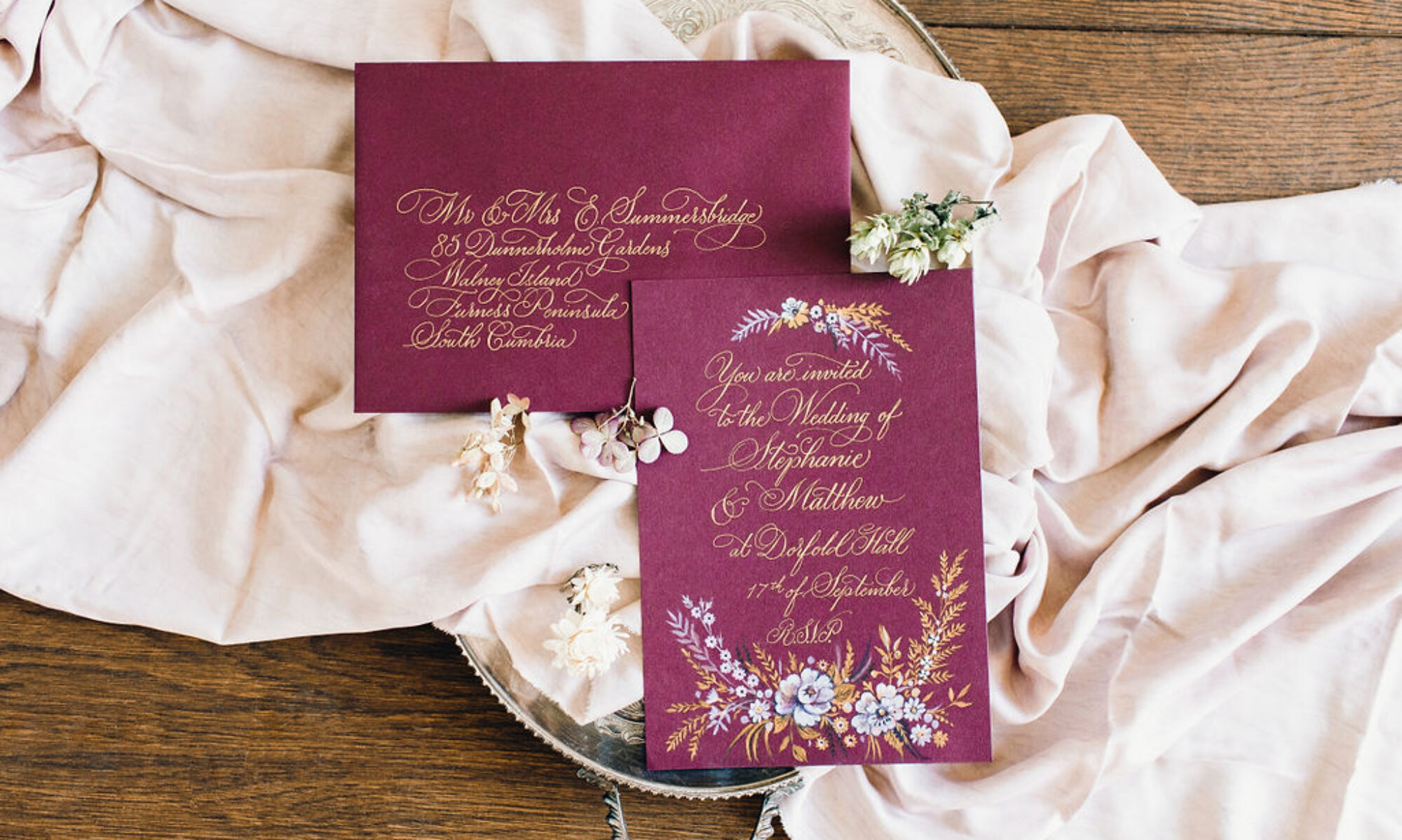 Swirly gold calligraphy on a burgundy wedding envelope and invitation. The invite is illustrated with white and gold flowers and leaves. The suite sits on scrunched up silk with hydrangea flowers and hops around it