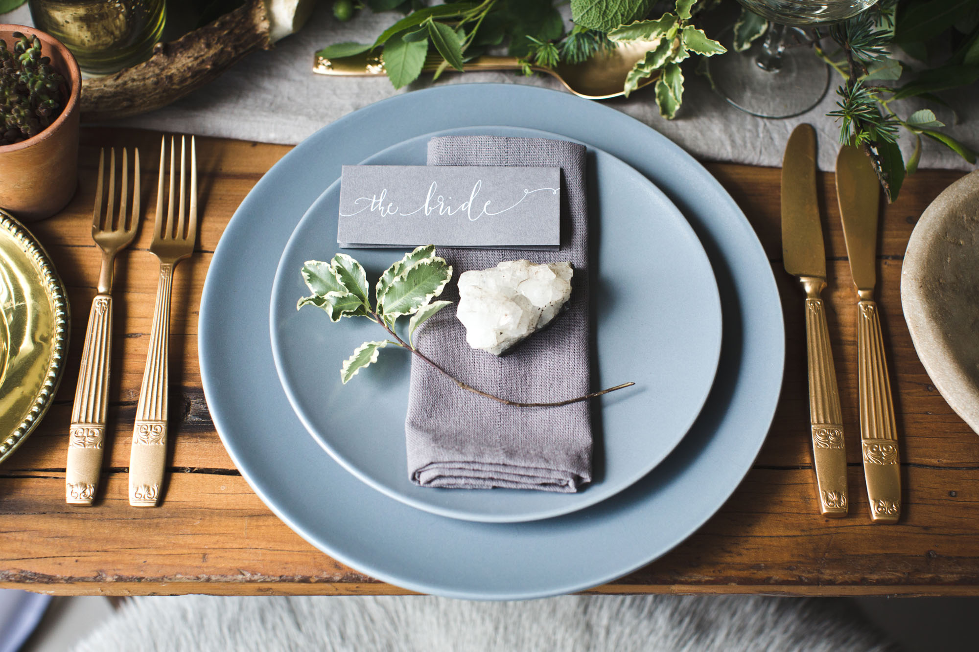 A grey place name with swirly calligraphy saying "the bride" sits on a grey napkin on dusky blue plates, with foliage around
