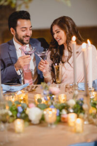 A couple clink glasses as they smile and chat. They're sat at a wedding table covered with candles, flowers and foliage