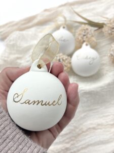 Hand holding a personalised Christmas bauble with Samuel written on it. The bauble is white and there are 2 more in the background