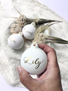 Three white Christmas tree decorations with with names in gold letters and a hand picking one up
