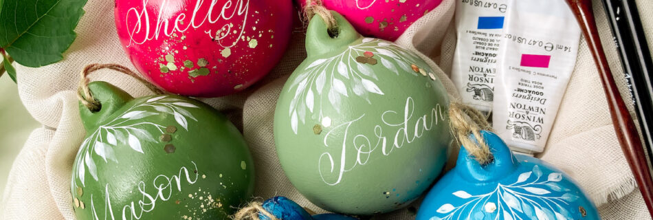 A bowl of colourful hand painted Christmas baubles with names on, next to some leaves and a calligraphy pen and paints