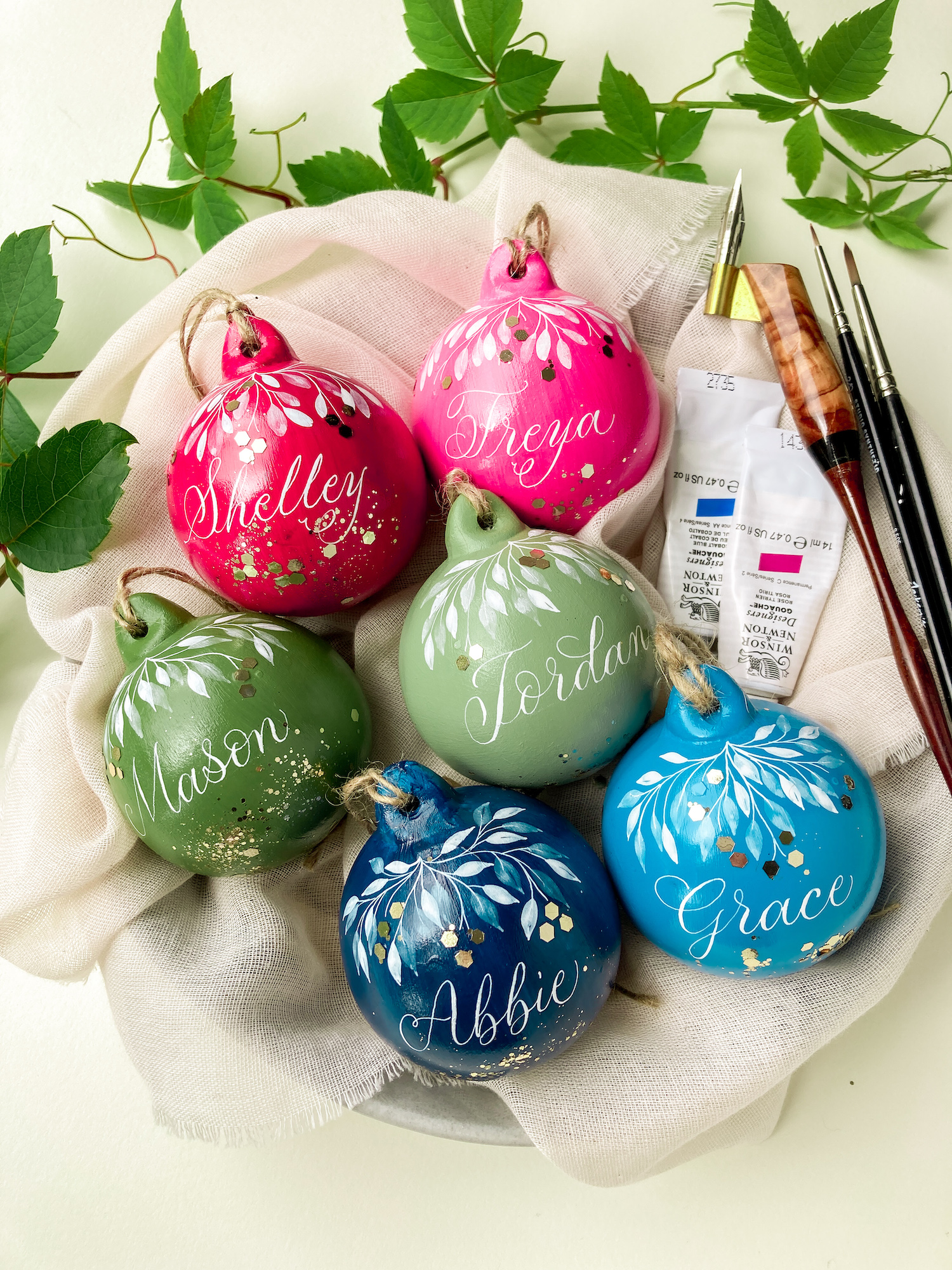 A bowl of colourful hand painted Christmas baubles with names on, next to some leaves and a calligraphy pen and paints